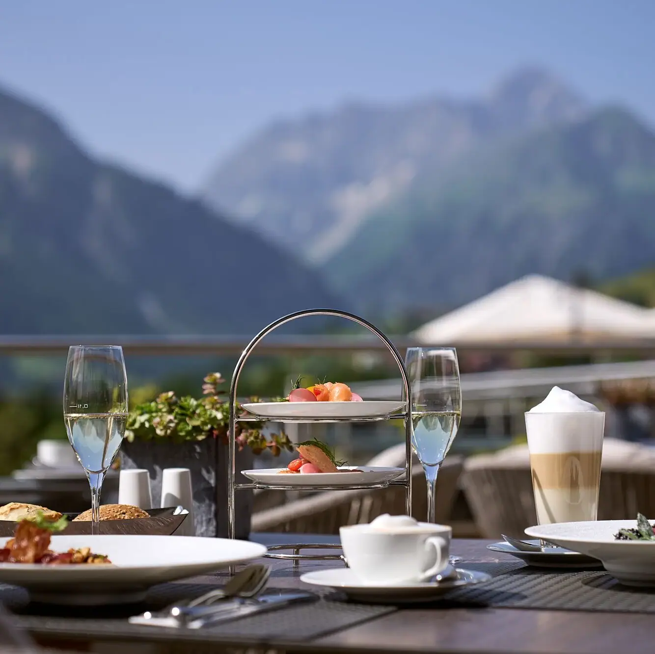 Ifen Hotel Kleinwalsertal Ein Tisch mit Tellern voller Speisen und Getränken vor einer Bergkulisse mit blauem Himmel..