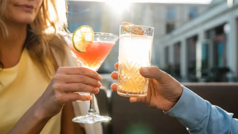 Cockatils Zwei Personen stoßen bei Sonnenuntergang auf einer Terrasse mit Getränken an. Im Fokus steht eine Hand, die einen Cocktail mit Limettenscheibe in einem Martini-Glas hält, und eine andere Hand, die ein Highball-Glas mit einem klaren, sprudelnden Getränk und Orangenscheibe hält. Die warme Abendsonne beleuchtet die Szene, was eine entspannte und gesellige Stimmung vermittelt.