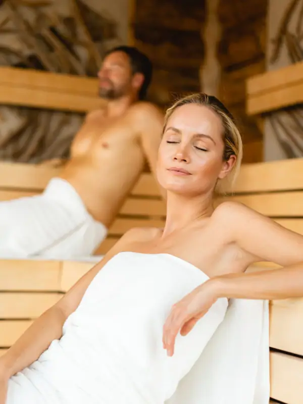 Eine Frau sitzt in einer Sauna, ein Mann hinter ihr.