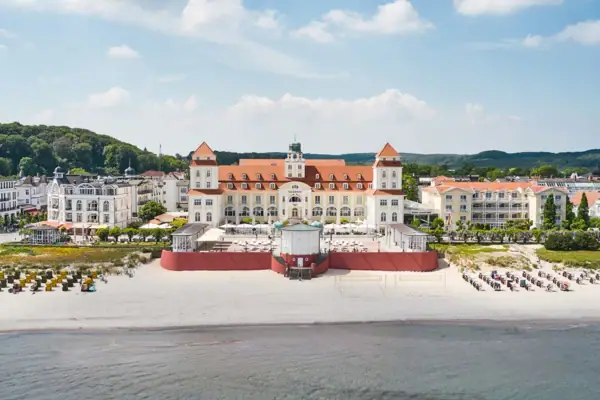 Kurhaus Binz Großes Gebäude am Strand mit einem Gewässer im Vordergrund.