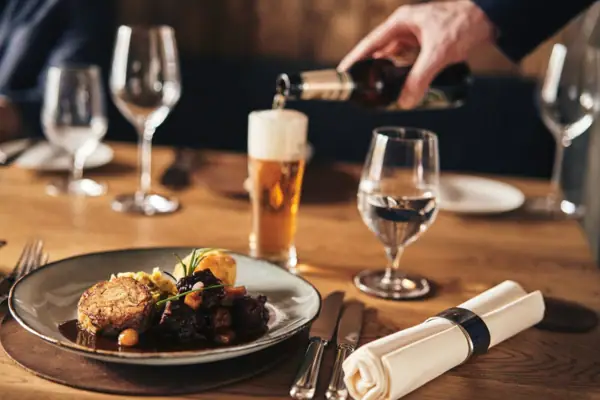 Walser Stube Carnozet im Ifen Hotel Kleinwalsertal Auf einem gedeckten Holztisch steht schön angerichtetes, deftiges Gericht auf einem Teller, während eine Hand im Hintergrund Bier in ein Glas eingießt.