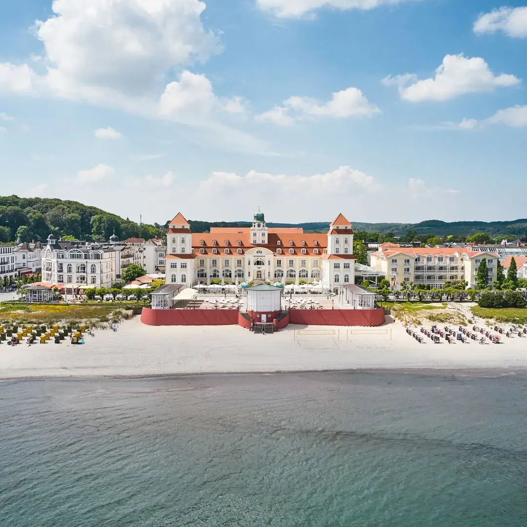 Kurhaus Binz Großes Gebäude am Strand mit einem Gewässer im Vordergrund.