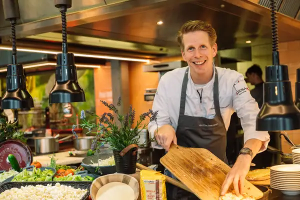Koch Travemünde Ein Koch mit Kochschürze steht in einer Küche, hinter dem Buffet und hält ein Schneidebrett in der Hand.
