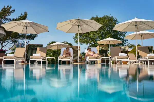 Eine Gruppe von Menschen sitzt auf Stühlen am Pool unter Sonnenschirmen.