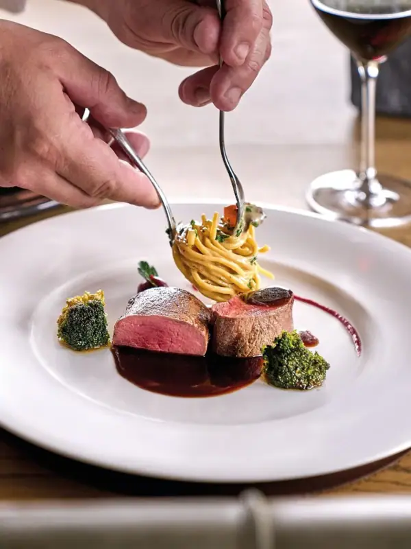 Eine Person hält eine Gabel beim Anrichten eines Gerichts über einem Teller mit Essen.