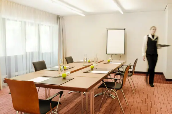 Dialog II Eine Frau steht in einem Tagungsraum mit einem Tischblock und Stühlen drumherum.