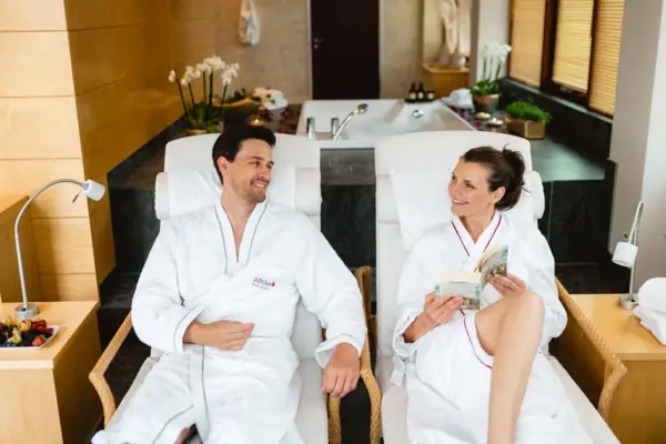 Spa Suite Travemünde Ein Mann und eine Frau in weißen Bademänteln liegen in einem gemütlichen SPA Bereich und entspannen sich, die Frau hält ein Buch in den Händen.