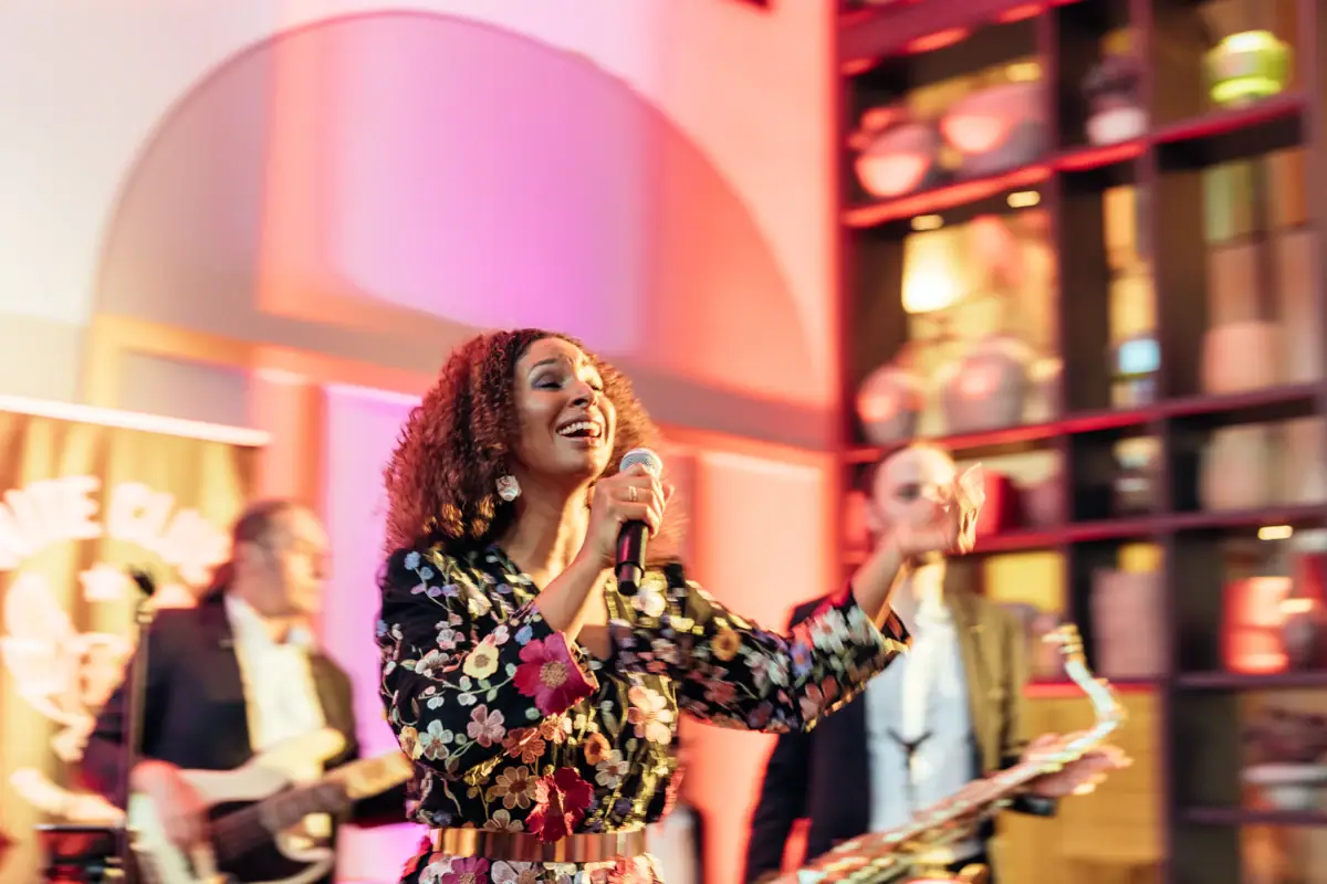 Frau mit lockigen Haaren und geblümtem Outfit singt fröhlich ins Mikrofon, begleitet von einer Live-Band mit Bassist und Saxophonist, in stimmungsvoll beleuchtetem Innenraum.