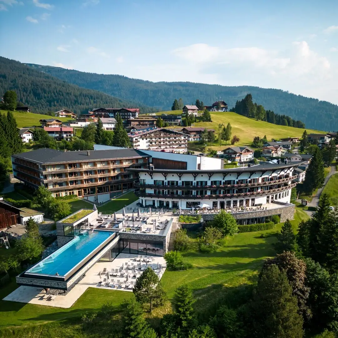 Angebote im Ifen Hotel Kleinwalsertal Ein Gebäude mit einem Infinity Pool inmitten eines Bergdorfs, umgeben von Bäumen und Grünflächen.