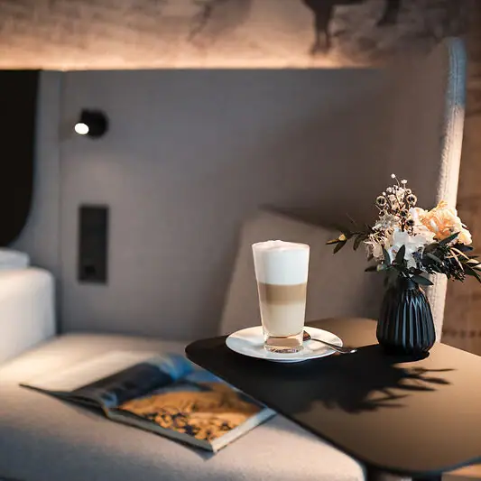 Zimmer & Suiten im Kleinwalsertal Ein Beistelltisch mit einem Latte Macchiato, einer aufgeschlagenen Zeitschrift und einer Vase mit Blumen neben einem Bett mit gepolstertem Kopfteil vor einer kunstvoll gestalteten Tapete..
