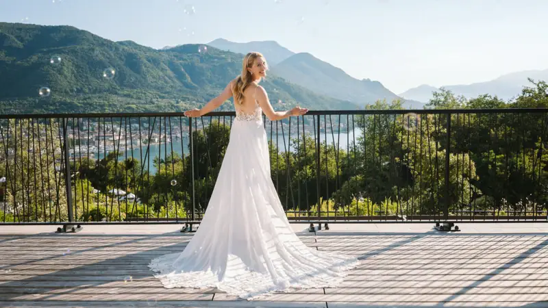 Heiraten im A-ROSA Gardasee Eine Frau in einem weißen Hochzeitskleid steht auf einer Terrasse mit Geländer und Bergen sowie dem Gardasee im Hintergrund.