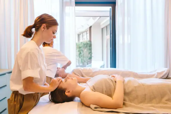 Eine Frau liegt auf dem Rücken und erhält von einer Masseurin eine Gesichtsmassage, während im Hintergrund eine Person auf einer zweiten Liege ebenfalls massiert wird.