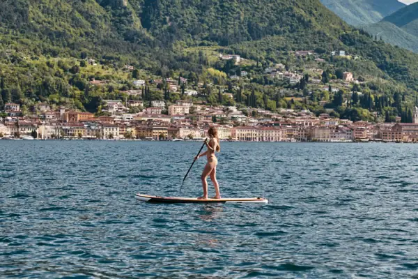 SUP Gardasee Eine Frau fährt mit einem Stand-up-Paddle Board auf dem Garadsee vpr der Kulisse einer Stadt vor grün bewachsenen Hängen an der Küste.