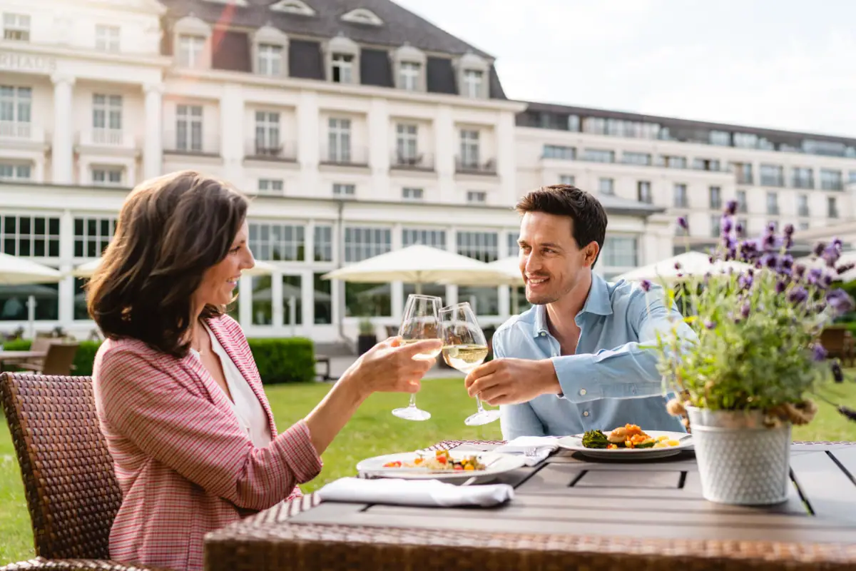 Terrasse A-ROSA Travemünde Ein Mann und eine Frau stoßen mit Weingläsern an einem Tisch im Freien an.