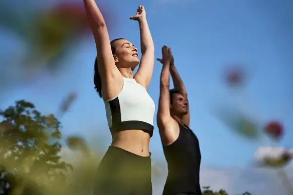 Yoga Eine Frau und ein Mann tehen im Freien uns halten die Arme neben den Ohren bei einer Yogaübung.