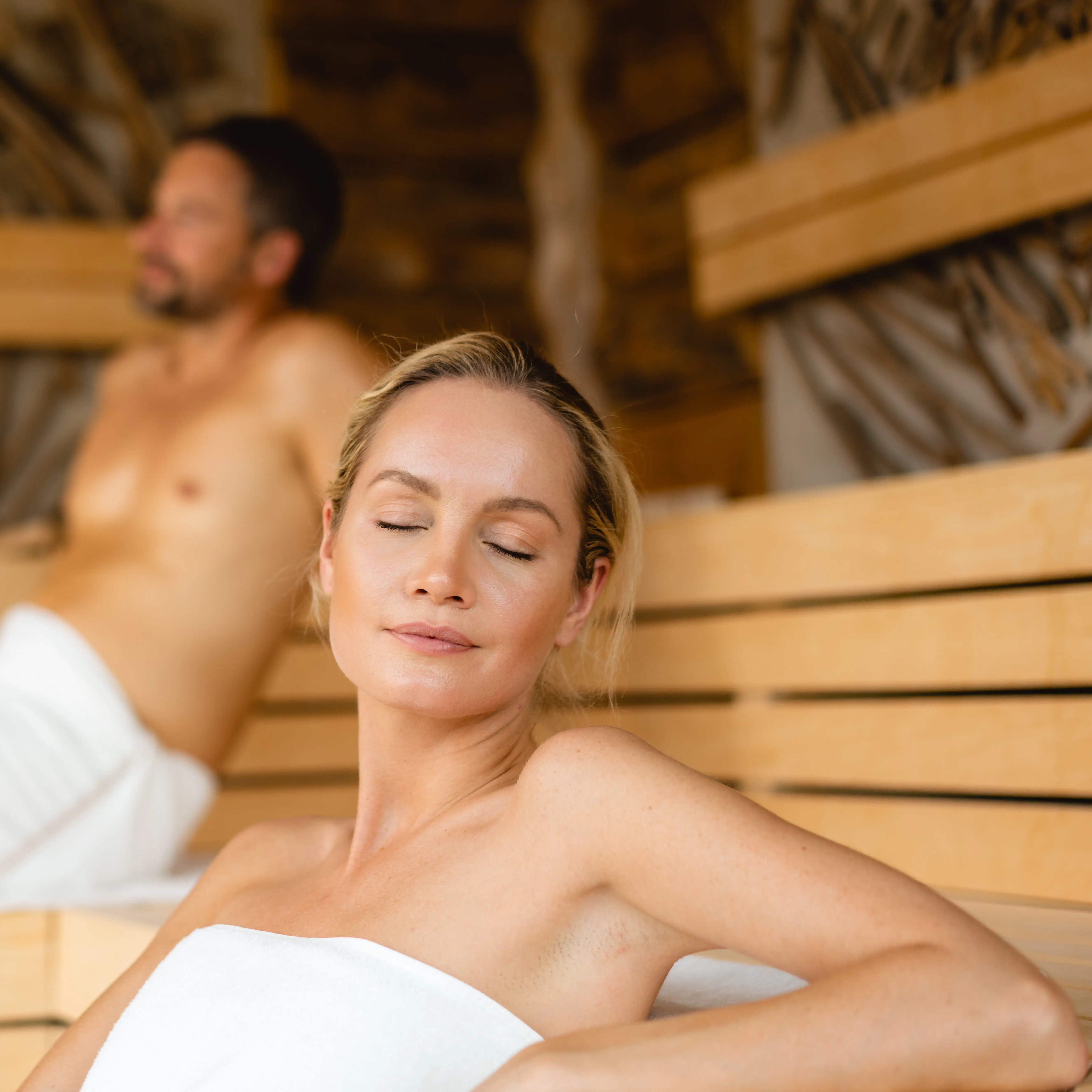 Eine Frau sitzt in einer Sauna, mit einem Mann im Hintergrund.