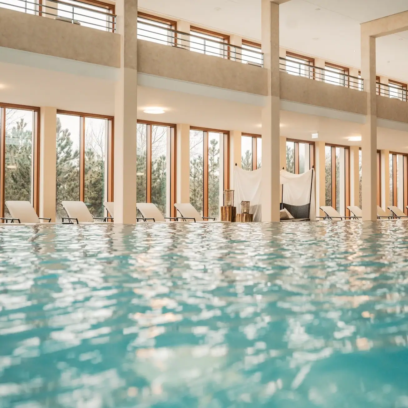 Innenpool im A-ROSA Sylt Ein Schwimmbecken mit Liegestühlen am Rand und Ausblick auf die Dünenlandschaft durch die Fenster im Hintergrund..