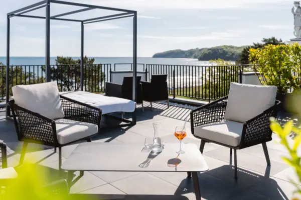 Day-Bed und Lounge-Sessel auf einer Dachterrasse in der Sonne mit dem Meer im Hintergrund.