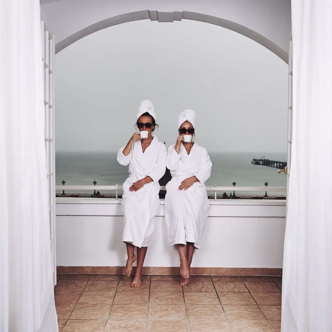 Day SPA Mädelstag Zwei Frauen in Bademänteln mit Sonnenbrillen und Kaffeetassen stehen auf einem Balkon