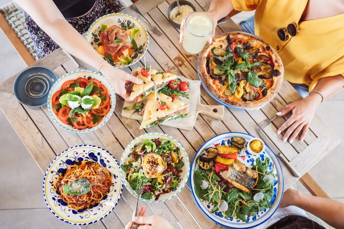 Kulinarik im A-ROSA Gardasee Eine Gruppe von Menschen isst gemeinsam an einem Tisch. Es gibt Pasta, Tomate Mozzarella, vegetairsche Pizza, Lachs mit Salat. Eine Frau nimmt sich gerade ein Focaccia.