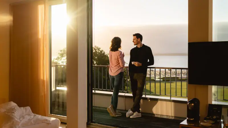 Zimmer & Suiten Ein Mann und eine Frau stehen auf einem Balkon und blicken aus einem Fenster.